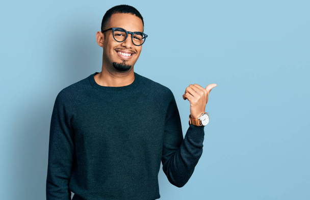 Joven hombre afroamericano vistiendo ropa casual y gafas sonriendo con la cara feliz mirando y señalando a un lado con el pulgar hacia arriba.  - Foto, Imagen