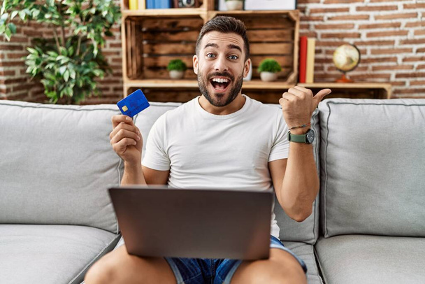 Красивый латиноамериканец, делающий покупки в интернете, держит кредитку, указывая пальцем вверх в сторону, улыбаясь счастливо с открытым ртом.  - Фото, изображение