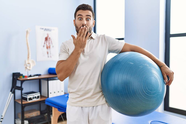 Bonito homem hispânico segurando bola pilates na clínica de reabilitação cobrindo a boca com a mão, chocado e com medo de erro. expressão surpreendida  - Foto, Imagem