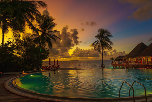 Luxury sunset over infinity pool in a summer beachfront hotel resort at tropical landscape. Tranquil beach holiday vacation background mood. Amazing island sunset beach view, palms swimming pool - Photo, Image