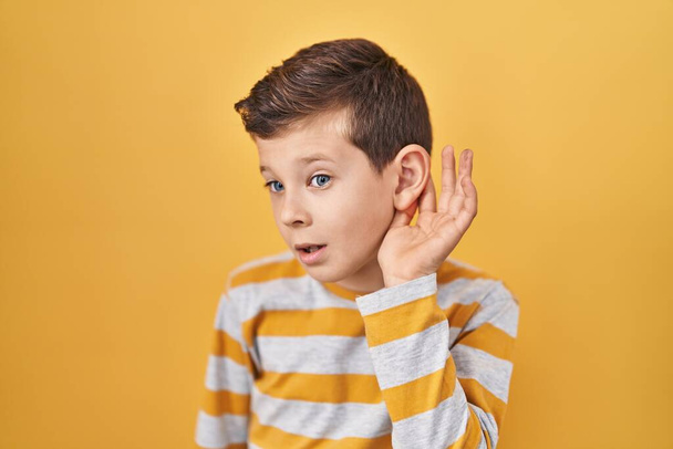 Młody biały dzieciak stojący nad żółtym tłem uśmiechający się z ręki na ucho słuchający plotek lub plotek. koncepcja głuchoty.  - Zdjęcie, obraz