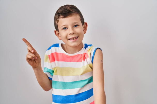 Молода кавказька дитина отримує вакцину, показуючи руку з груповою допомогою посміхаючись щасливим вказівником рукою і пальцем на бік
  - Фото, зображення