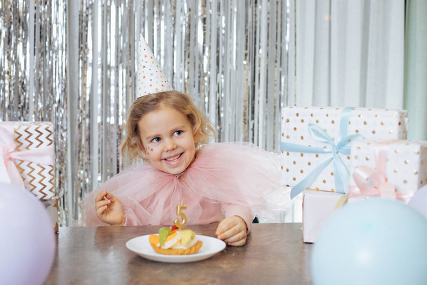 Childrens positive emotions. Happy smiling girl in festive outfit and party hat is sitting in front of birthday cake decorated with number five, surrounded by many gift boxes and balloons. - Foto, imagen