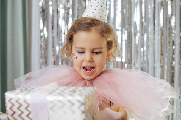 Святкування дня народження. Щаслива святкова одягнена 4-5-річна дівчина з зірками на щоках у вечірньому капелюсі тримає подарункову коробку в руках. Подарунки дітям
. - Фото, зображення