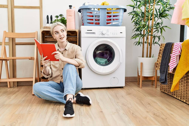 Молода біла жінка читає книгу в очікуванні пральної машини в пральні
 - Фото, зображення