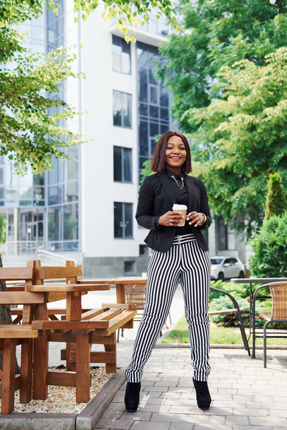 Coffee break. Young afro american woman in fashionable clothes outdoors in the city near green trees and against business building. - Foto, afbeelding
