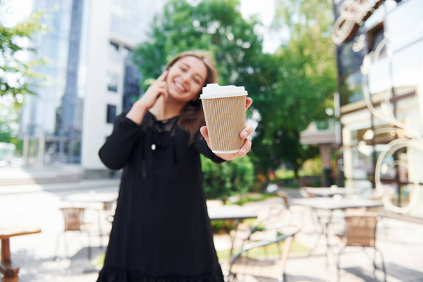 Talking by phone and holding cup of drink. Happy young girl in black skirt outdoors in the city near green trees and against business building. - Фото, зображення