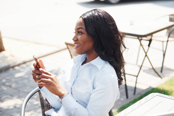 Sitting by table with phone in hands. Young afro american woman in white shirt outdoors in the city. - Foto, imagen