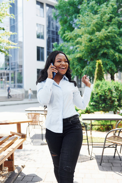 Having conversation by using phone. Young afro american woman in white shirt outdoors in the city near green trees and against business building. - Фото, изображение