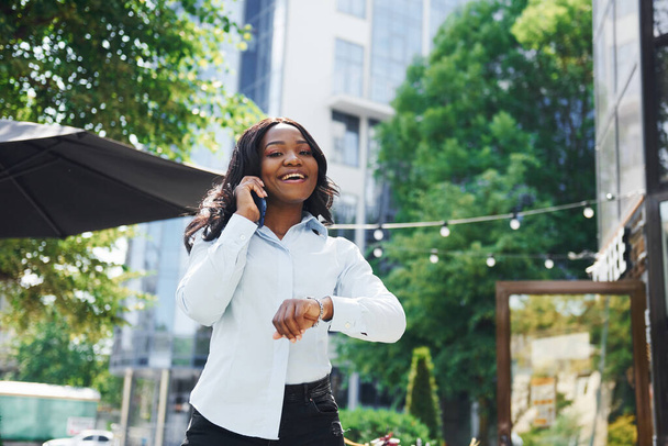 Having conversation by using phone. Young afro american woman in white shirt outdoors in the city near green trees and against business building. - Foto, afbeelding