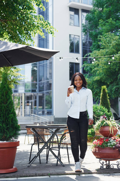 Holding cup of drink in hands. Young afro american woman in white shirt outdoors in the city near green trees and against business building. - Photo, image