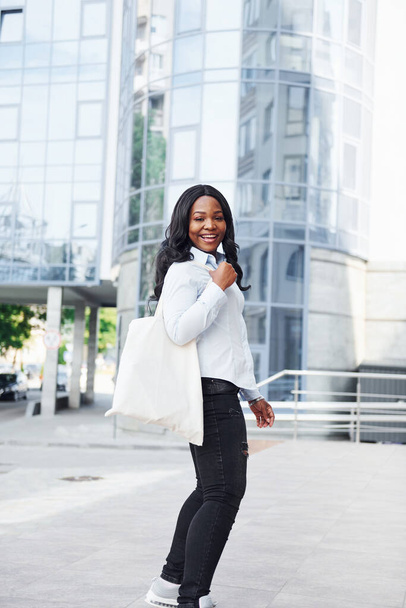 With shopping bag. Young afro american woman in white shirt outdoors in the city against business building. - Photo, image