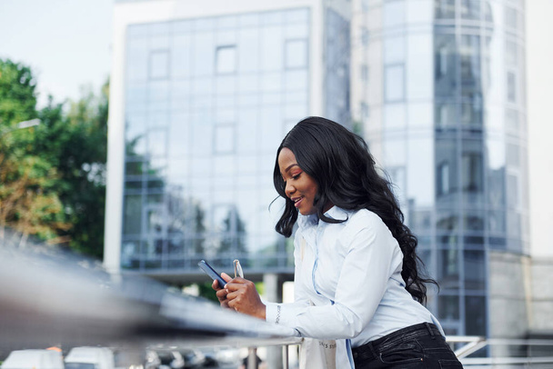 With phone in hands. Young afro american woman in white shirt outdoors in the city against business building. - Fotoğraf, Görsel