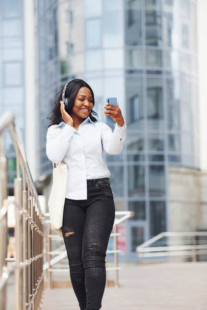 Ik luister naar muziek in een koptelefoon. jong afro amerikaanse vrouw in wit shirt buiten in de stad tegen business building. - Foto, afbeelding