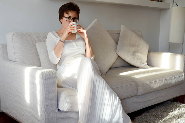 Beyaz etekli, rahat gömlekli, rahat rahat kanepede oturan mutlu bir kadının hayat tarzı portresi. Kahve içiyorum. Hayal kurmak. - Fotoğraf, Görsel