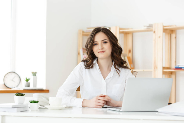 Молодая деловая женщина или секретарь, сидящая за столом и работающая. Улыбаясь и глядя в камеру - Фото, изображение