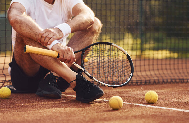 Sitting and taking a break. Close up view of senior man in white shirt and black sportive shorts that is on tennis court. - Φωτογραφία, εικόνα