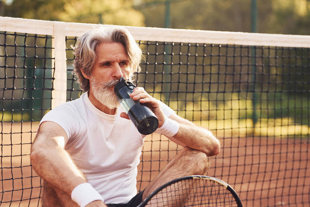 Taking break and drinking water. Senior stylish man in white shirt and black sportive shorts on tennis court. - Photo, image