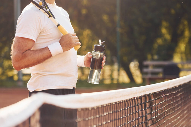 Taking break and drinking water. Senior stylish man in white shirt and black sportive shorts on tennis court. - Фото, зображення