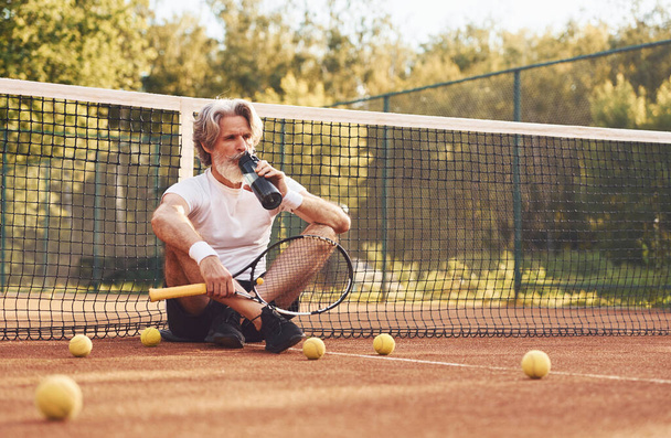 Taking break and drinking water. Senior stylish man in white shirt and black sportive shorts on tennis court. - Foto, afbeelding