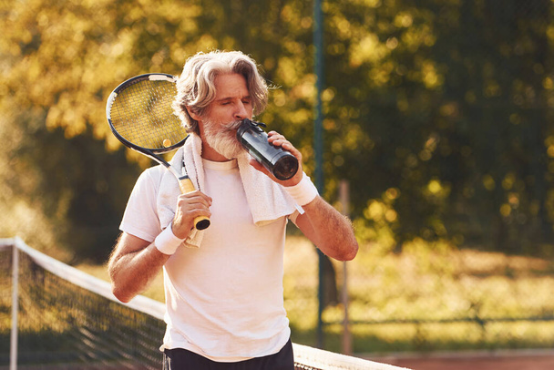 Taking break and drinking water. Senior stylish man in white shirt and black sportive shorts on tennis court. - Photo, image