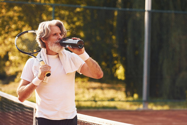 Taking break and drinking water. Senior stylish man in white shirt and black sportive shorts on tennis court. - Foto, immagini