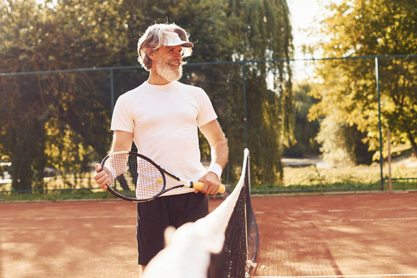 Beautiful sunlight. Senior stylish man in white shirt and black sportive shorts on tennis court. - Foto, imagen