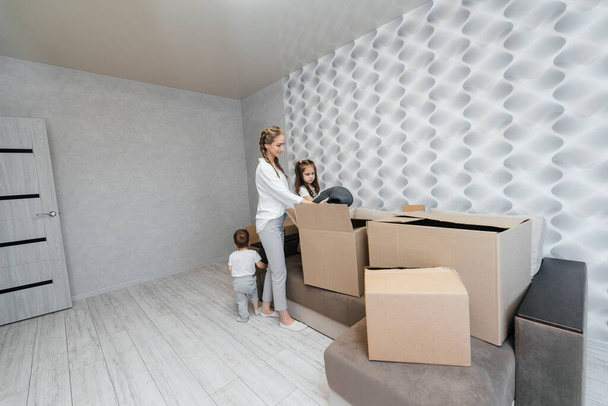 A young mother with children unpacks a box of things after moving to a new apartment. Purchase of real estate. Housewarming, delivery and transportation of goods. - Фото, изображение