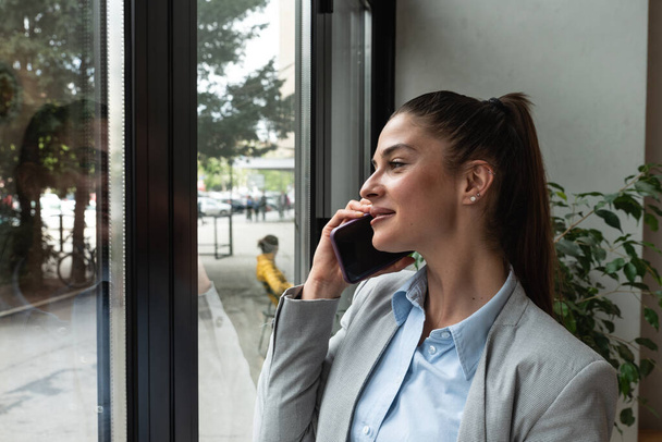 Young business woman in casual suit standing near window in the restaurant holding her smartphone waiting to waiter pack her lunch to go in the box. Freelancer expat waiting take away coffee or food. - Fotó, kép