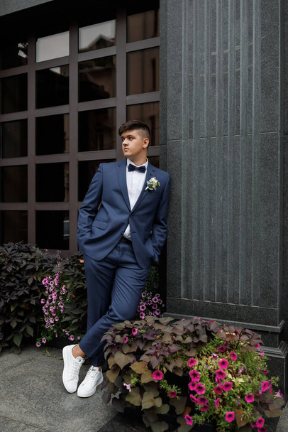 Een jonge bruidegom in een blauw pak en witte sneakers. Een prachtig gebouw op de achtergrond. - Foto, afbeelding