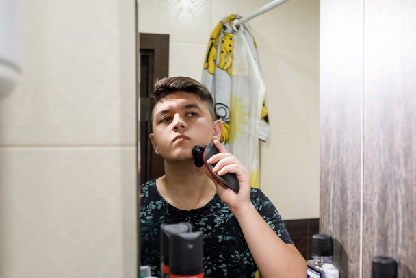 Ένας άντρας ξυρίζεται με ένα ηλεκτρικό ξυράφι στο μπάνιο, μια αντανάκλαση στον καθρέφτη.. - Φωτογραφία, εικόνα