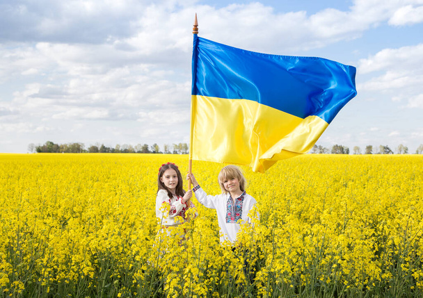 ウクライナの民族衣装の男の子と女の子は、黄色の菜の花畑の中に黄色の青い旗を立って保持しています。愛国心の教育。独立記念日。ウクライナの子供たちは平和を求めて - 写真・画像