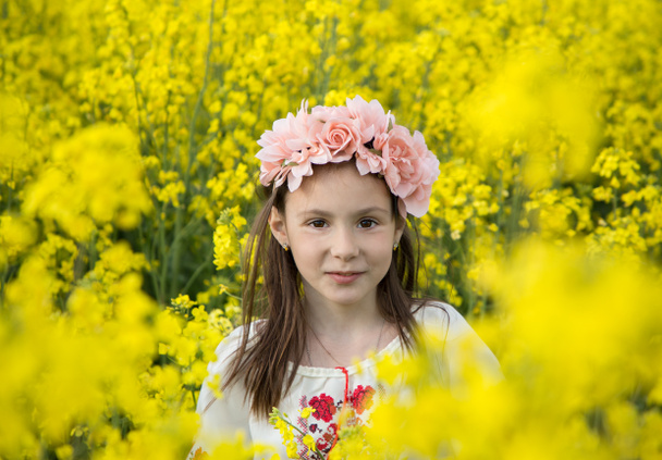 Ritratto di una ragazza carina di 7 anni in una ghirlanda e una tradizionale camicetta ricamata tra un campo di colza giallo fiorito. Bambini per la pace. Stai con l'Ucraina. Sii orgoglioso di essere ucraino - Foto, immagini