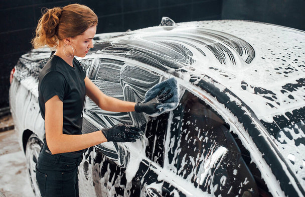 Wipes vehicle that is in white soap. Modern black automobile get cleaned by woman inside of car wash station. - Fotoğraf, Görsel