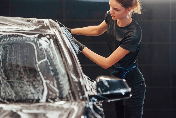 Wipes vehicle that is in white soap. Modern black automobile get cleaned by woman inside of car wash station. - Фото, изображение