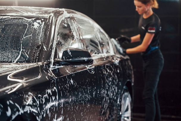 Wipes vehicle that is in white soap. Modern black automobile get cleaned by woman inside of car wash station. - Foto, imagen