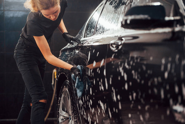 Wipes vehicle that is in white soap. Modern black automobile get cleaned by woman inside of car wash station. - Photo, image