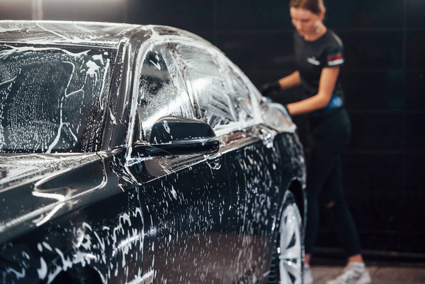 Wipes vehicle that is in white soap. Modern black automobile get cleaned by woman inside of car wash station. - Foto, Bild