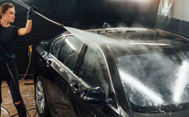 Using equipment with high pressure water. Modern black automobile get cleaned by woman inside of car wash station. - Fotoğraf, Görsel