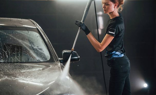 Using equipment with high pressure water. Modern black automobile get cleaned by woman inside of car wash station. - Zdjęcie, obraz