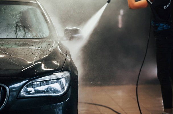 Using equipment with high pressure water. Modern black automobile get cleaned by woman inside of car wash station. - Фото, изображение