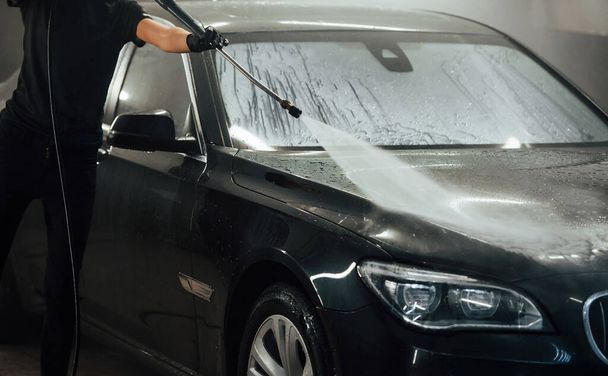 Using equipment with high pressure water. Modern black automobile get cleaned by woman inside of car wash station. - 写真・画像