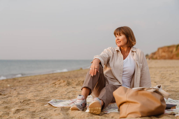 Senior european woman smiling while resting on sandy beach - Foto, afbeelding