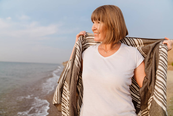 Senior european woman smiling while walking on sandy beach - Foto, afbeelding