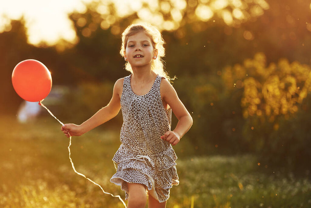 Happy little girl with red balloon in hands have fun outdoors on meadow at summertime. - Foto, imagen