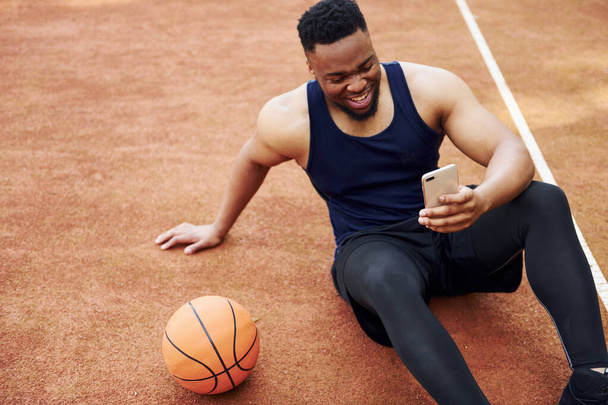 Using phone. African american man plays basketball on the court outdoors. - Φωτογραφία, εικόνα