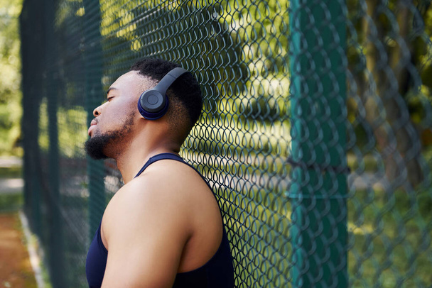 Afrikai amerikai fickó vezeték nélküli fülhallgatóval támaszkodik a sportpálya fémhálójára, és szünetet tart.. - Fotó, kép