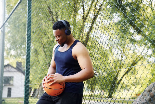 African american man with wireless headphones takes a break and leaning on the metal mesh with ball on the court outdoors. - Фото, изображение