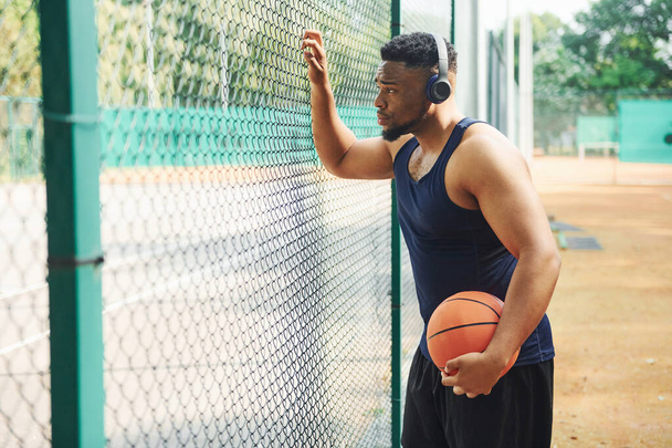 African american man with wireless headphones takes a break and leaning on the metal mesh with ball on the court outdoors. - Foto, immagini