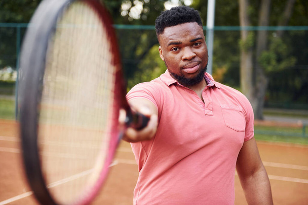 African american man in pink shirt posing for a camera with tennis racket on the court outdoors. - Valokuva, kuva